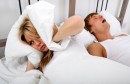 Woman covering ears with pillow while man snoring in bed